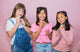 Three young girls posing together against a pink background
