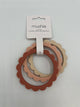 The rose, blush, and sifting sand Mushie Flower Teething Bracelets in the package. It shows the 3 rings (from left to right rose (dusty pink), blush (light pink), sifting sand (cream). They are all held together by a white paper label that reads, "mushie, silicone, flower teething bracelet."