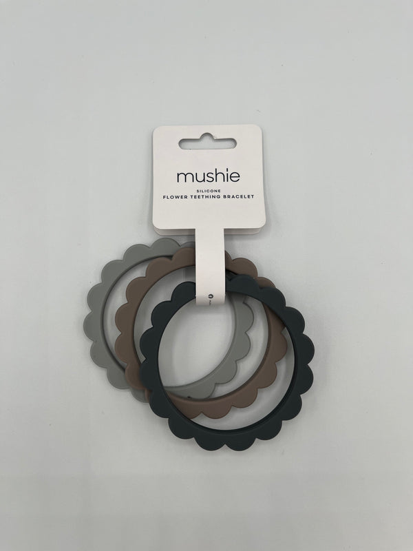 The steel, dove, and stone Mushie Flower Teething Bracelets in the package. It shows the 3 rings (from left to right dove (light grey), stone (taupe), stone (dark grey)). They are all held together by a white paper label that reads, "mushie, silicone, flower teething bracelet."