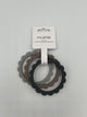 The steel, dove, and stone Mushie Flower Teething Bracelets in the package. It shows the 3 rings (from left to right dove (light grey), stone (taupe), stone (dark grey)). They are all held together by a white paper label that reads, "mushie, silicone, flower teething bracelet."