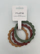 The dried thyme, berry, and natural Mushie Flower Teething Bracelets in the package. It shows the 3 rings (from left to right natural (brown), berry (maroon), dried thyme (sage green)). They are all held together by a white paper label that reads, "mushie, silicone, flower teething bracelet."