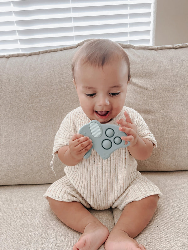 Baby sitting on a tan couch in a tan outfit holding the grey silicone elephant Itzy Pop.