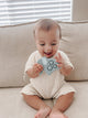 Baby sitting on a tan couch in a tan outfit holding the grey silicone elephant Itzy Pop.