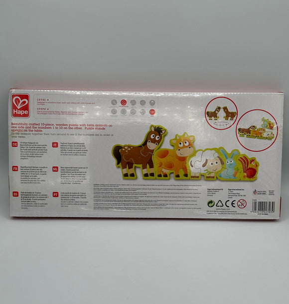 Back of the Hape Numbers and Farm Animals box. It is white with a picture of the puzzle on it. The puzzle has a light green border with a horse, cow, sheep, rabbit, and rooster in a line. In the top right corner there are two circles, one shows the front of the horse piece and the back of the horse piece that contains numbers, and the other show the pieces of the puzzle.
