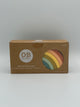 The silicone rainbow stacker in the package. The package is a cardboard box that says, "OB Designs, Rainbow Stacker, Designed with love in Byron Bay, Australia." On the right side is a clear circle cutout through which you can see the silicone stacker that is (from outside to inside) red, pink, yellow, olive green, mint green, and teal with a small visible piece of the yellow inside.