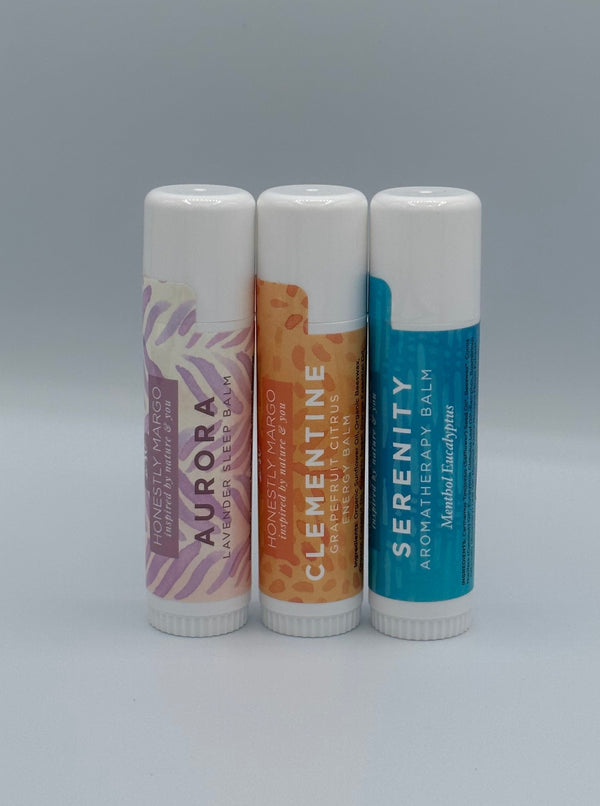 The three Honestly Margo Aromatherapy Balms. (From left to right) There is a white tube with purple leaves that says the scent, "Aurora, Lavender Sleep Balm" on it, an orange tube with orange flowers that says, "Clementine. Grapefruit Citrus Energy Balm", and a blue tube that says, "Serenity, Aromatherapy Balm".