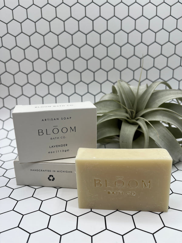 The Lavender Bloom body bar. The soap is in the front and says, "Bloom Bath Co", and the box is in the back stacked on top of a laying down box. It is white and says "Bloom Bath Co Lavender". There is an air plant in the back corner.