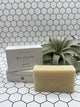 The Lavender Bloom body bar. The soap is in the front and says, "Bloom Bath Co", and the box is in the back stacked on top of a laying down box. It is white and says "Bloom Bath Co Lavender". There is an air plant in the back corner.