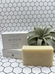 The eucalyptus Bloom body bar. The soap is in the front and says, "Bloom Bath Co", and the box is in the back stacked on top of a laying down box. It is white and says "Bloom Bath Co Eucalyptus". There is an air plant in the back corner.