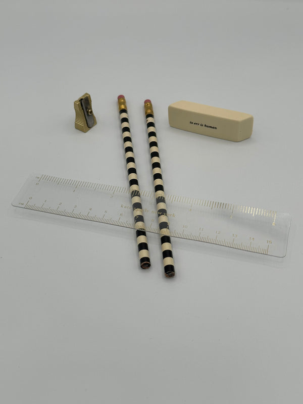Small gold pencil sharpener, two tan and black striped pencils, a tan eraser, and a clear ruler with gold markings.