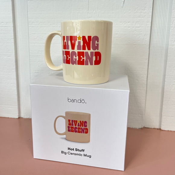 The Ban-do Hot Stuff mug on top of the box. The mug is cream and says, "Living Legend" with purple and red writing. The box is white with a photo of the mug on it and says, "Hot stuff big ceramic mug".