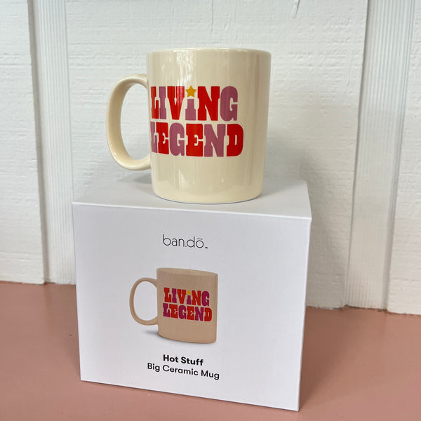 The Ban-do Hot Stuff mug on top of the box. The mug is cream and says, "Living Legend" with purple and red writing. The box is white with a photo of the mug on it and says, "Hot stuff big ceramic mug".