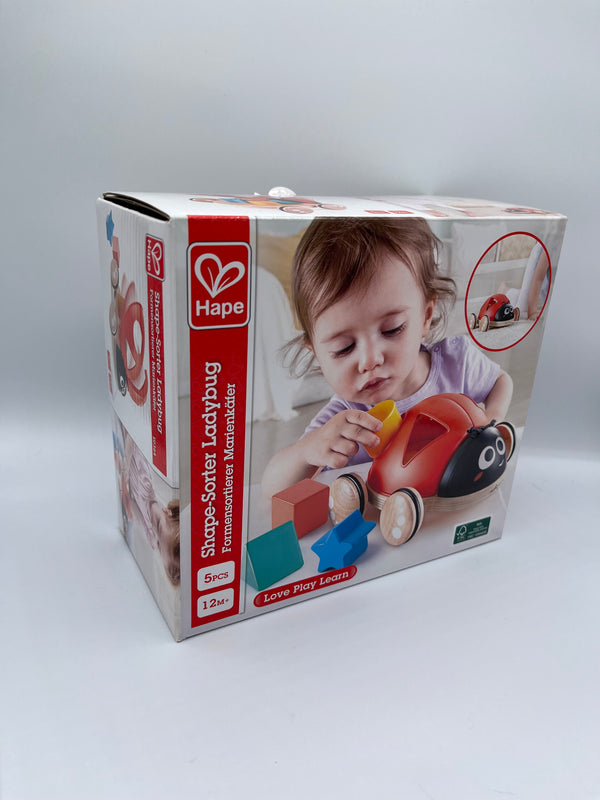 Hape Shape Sorter Ladybug box. There is an image of a ladybug with red wings and cutouts of a triangle, circle, square, and star with 4 wooden wheels. In front there is a 3d green triangle, blue star, and orange square. There is a little girl putting the 3d yellow circle through the circle cutout.  Along the left side of the box there is a red strip that says, "Shape-Sorter Ladybug, 5pcs, 12M+".
