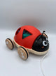 Front of the Hape Shape Sorter Ladybug. The red wings with a circle, triangle, star, and square are closed. Through the triangle you can see the 3d blue star and green triangle sitting on the wooden platform. The ladybug has a black head with two eyes and two antennas. There are 4 wooden wheels on the platform and a white string to pull the ladybug.