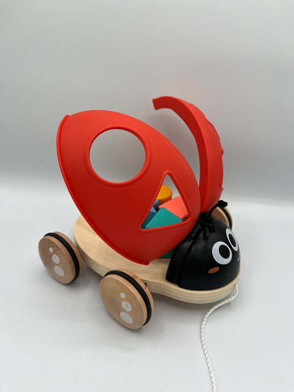 Front of the Hape Shape Sorter Ladybug. The red wings with a circle, triangle, star, and square are open. Through the triangle you can see the 3d yellow circle, blue star, green triangle, and orange square sitting on the wooden platform. The ladybug has a black head with two eyes and two antennas. There are 4 wooden wheels on the platform and a white string to pull the ladybug. 
