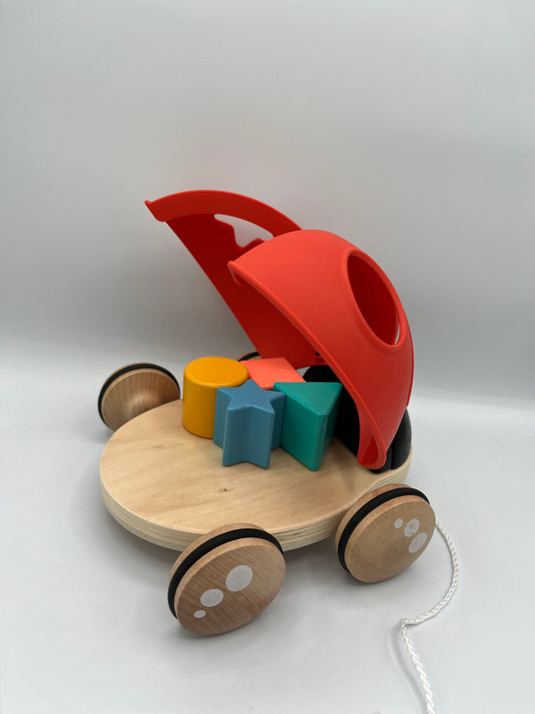 Back of the Hape Shape Sorter Ladybug. There is a wooden platform with 4 wooden wheels. On the platform there is a 3d yellow circle, orange square, blue star, and green triangle. The red wings to the ladybug are open facing upward. There is a white string to pull the ladybug.