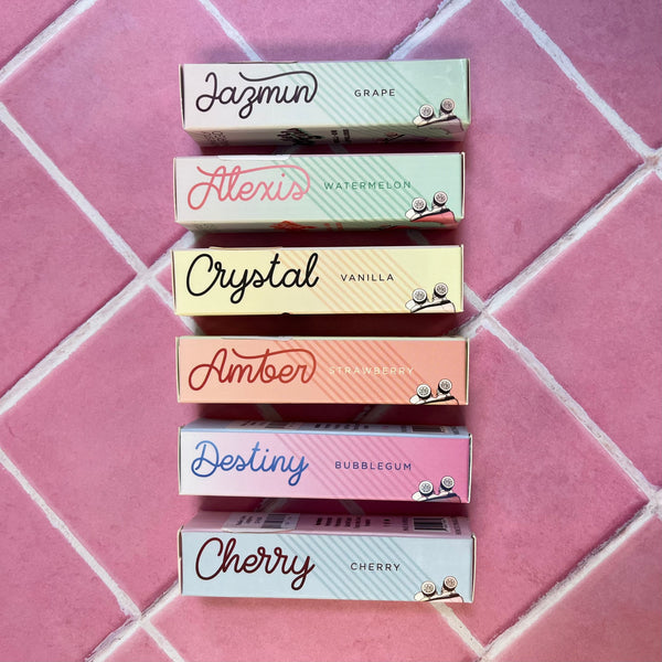 The sides of all 6 of the Honestly Margo Roller Girls Lip Glosses. (From top to bottom) There is a light mint green box that says, "Jazmin, grape" in black, a darker mint green box that says, "Alexis, watermelon" in pink, a yellow box that says, "Crystal, Vanilla" in black, a peach box that says, "Amber. Strawberry" in dark orange, a pink box that says, "Destiny, Bubblegum" in blue, and a light blue box that says, "Cherry, Cherry" in maroon.