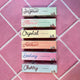 The sides of all 6 of the Honestly Margo Roller Girls Lip Glosses. (From top to bottom) There is a light mint green box that says, "Jazmin, grape" in black, a darker mint green box that says, "Alexis, watermelon" in pink, a yellow box that says, "Crystal, Vanilla" in black, a peach box that says, "Amber. Strawberry" in dark orange, a pink box that says, "Destiny, Bubblegum" in blue, and a light blue box that says, "Cherry, Cherry" in maroon.