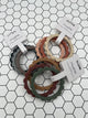 3 packs of the Mushie Flower Teething Bracelets. Each pack has 3 flower shaped rings. The pack in the back left has a navy, grey, and taupe ring. The pack in the back right has a light pink, rust, and tan ring. The pack in the front has a brown, maroon, and sage green ring.