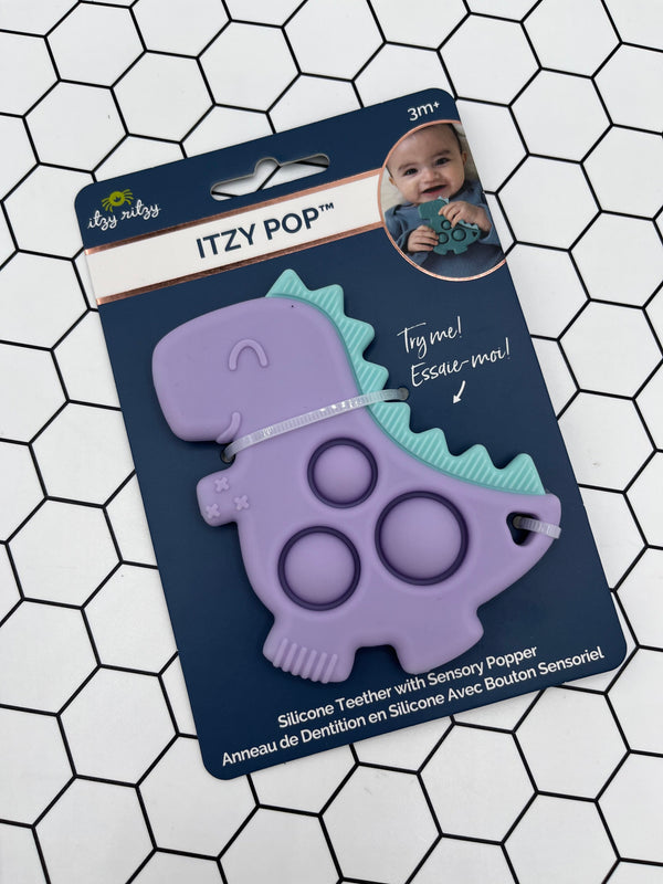 Purple dinosaur Itzy Pop in the packaging. The package is blue and says, "Itzy Pop". In the top right corner there is an image of a baby holding the teal dinosaur Itzy Pop. The product is a purple dinosaur with teal spikes and 3 popping bubbles in the middle. Along the bottom, it says, "Silicone Teether with Sensory Popper."
