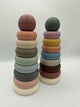 The original (left) and rustic (right) Mushie Stacking Rings. Each tower contains 8 rings of descending sizes going up and a ball on top. The original tower (from bottom to top) has a white, light pink, sage green, light blue, mint green, taupe, cream, and peach ring with a dusty blush pink ball on top. The rustic tower (from bottom to top) has  a white, dusty pink, brown, light grey, light blue, light orange, yellow, and light pink ring with a sage green ball on top.