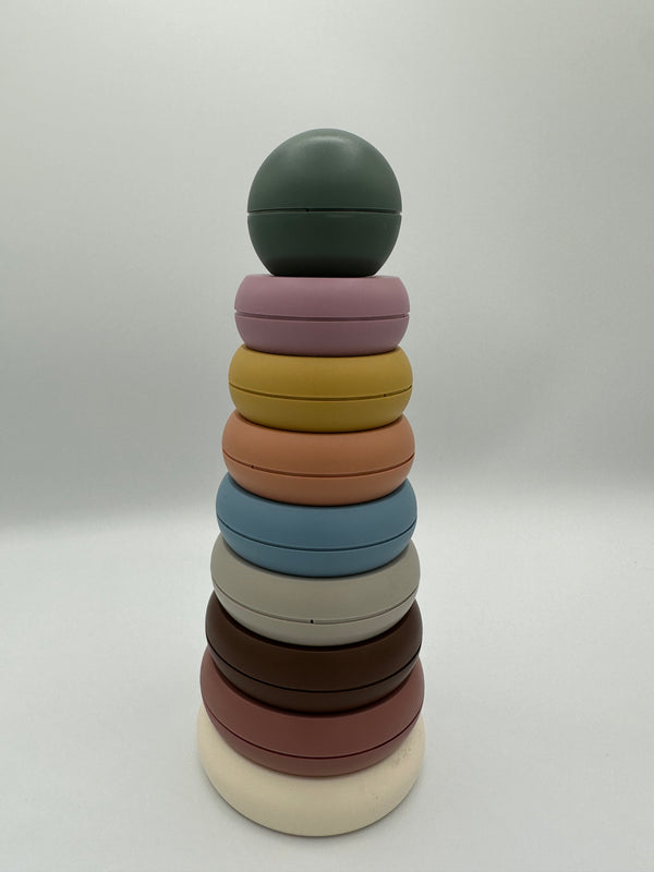 The rustic Mushie Stacking Rings tower. There are 7 rings of descending sizes going up to a ball on the top. The rings (from bottom to top) are white, dusty pink, brown, light grey, light blue, peach, yellow, and light purple with a sage green ball on top.