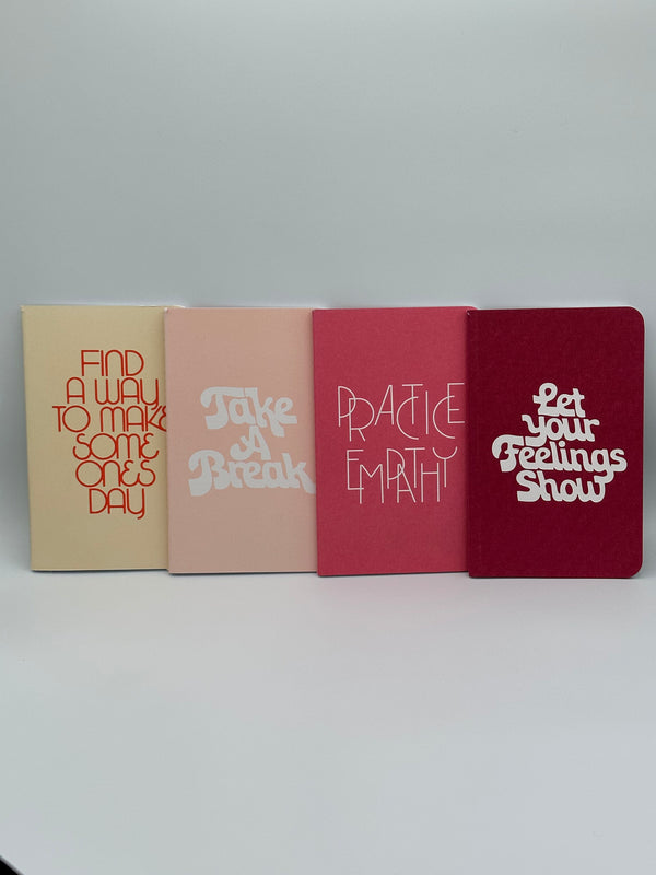 The cream, light pink, dark pink, and maroon notebooks in the Ban-do Wellness Notebook Set. The cream says, "Find a way to make someone's day" in red writing, the light pink says, "Take a break,'" the dark pink says, "practice empathy," and the maroon says, "Let your feelings show" in white writing.