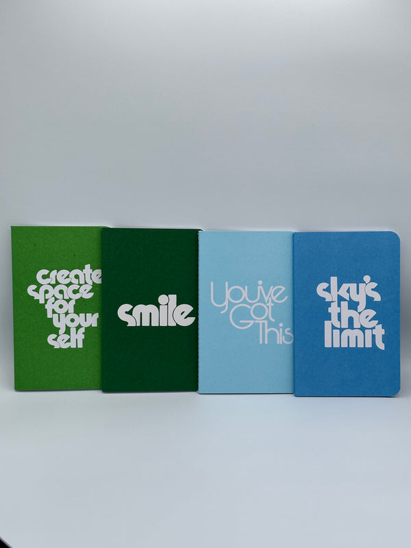 The dark green, light green, light blue, and dark blue notebooks in the Ban-do Wellness Notebook set. The light green says, "Create space for yourself," the dark green says, "smile," the light blue says, "You've got this," and the dark blue says, "sky's the limit."