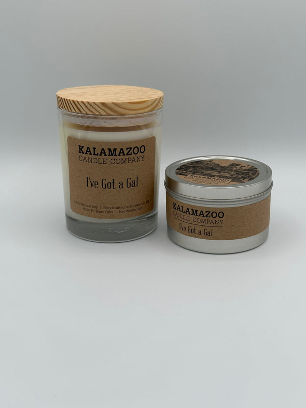 The I've Got a Gal Kalamazoo Candle jar next to the I've Got a Gal Kalamazoo Candle tin. The jar is clear through which you can see the white candle with a light wood lid and a brown paper label that reads, "Kalamazoo Candle Company, I've Got a Gal". The tin is a small cylindrical metal tin with a brown paper label that says, "Kalamazoo Candle Company, I've Got a Gal".