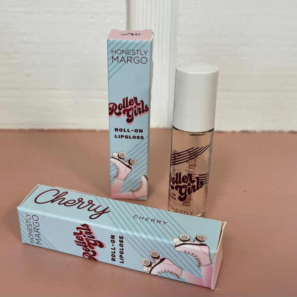 In the back there is the blue Honestly Margo Roller Girls Lip Gloss box next to the lip gloss. The box is blue and says, "Roller Girls, Roll-on Lipgloss", and the lip gloss is in a clear tube with a white cap and says, "Roller Girls" in maroon. In the front the box is laying on its side. On the top it says, "Cherry, Cherry", and on the side it says, "Roller Girls, Roll-on Lipgloss".