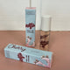 In the back there is the blue Honestly Margo Roller Girls Lip Gloss box next to the lip gloss. The box is blue and says, "Roller Girls, Roll-on Lipgloss", and the lip gloss is in a clear tube with a white cap and says, "Roller Girls" in maroon. In the front the box is laying on its side. On the top it says, "Cherry, Cherry", and on the side it says, "Roller Girls, Roll-on Lipgloss".