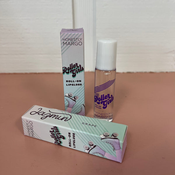 In the back there is the lavender Honestly Margo Roller Girls Lip Gloss box next to the lip gloss. The box is lavender and says, "Roller Girls, Roll-on Lipgloss", and the lip gloss is in a clear tube with a white cap and says, "Roller Girls" in purple. In the front the box is laying on its side. On the top it says, "Jazmin, Grape", and on the side it says, "Roller Girls, Roll-on Lipgloss".