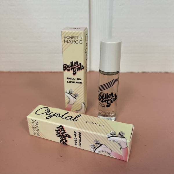 In the back there is the yellow Honestly Margo Roller Girls Lip Gloss box next to the lip gloss. The box is yellow and says, "Roller Girls, Roll-on Lipgloss", and the lip gloss is in a clear tube with a white cap and says, "Roller Girls" in black. In the front the box is laying on its side. On the top it says, "Crystal, Vanilla", and on the side it says, "Roller Girls, Roll-on Lipgloss".