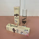 In the back there is the yellow Honestly Margo Roller Girls Lip Gloss box next to the lip gloss. The box is yellow and says, "Roller Girls, Roll-on Lipgloss", and the lip gloss is in a clear tube with a white cap and says, "Roller Girls" in black. In the front the box is laying on its side. On the top it says, "Crystal, Vanilla", and on the side it says, "Roller Girls, Roll-on Lipgloss".