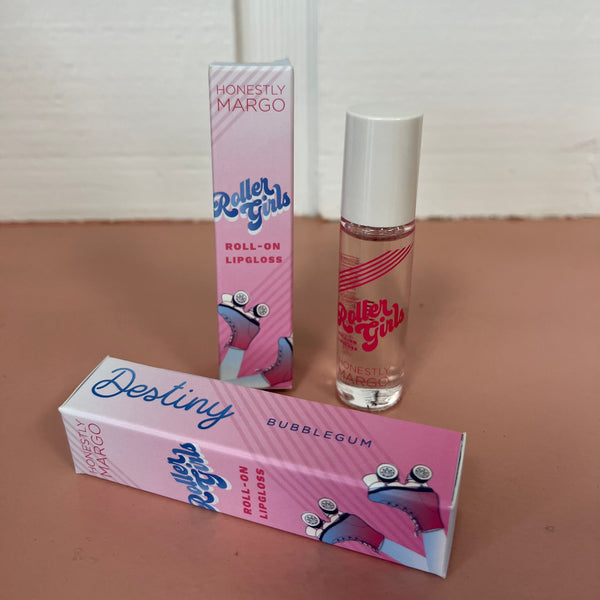 In the back there is the pink Honestly Margo Roller Girls Lip Gloss box next to the lip gloss. The box is pink and says, "Roller Girls, Roll-on Lipgloss", and the lip gloss is in a clear tube with a white cap and says, "Roller Girls" in pink. In the front the box is laying on its side. On the top it says, "Destiny, Bubblegum", and on the side it says, "Roller Girls, Roll-on Lipgloss".
