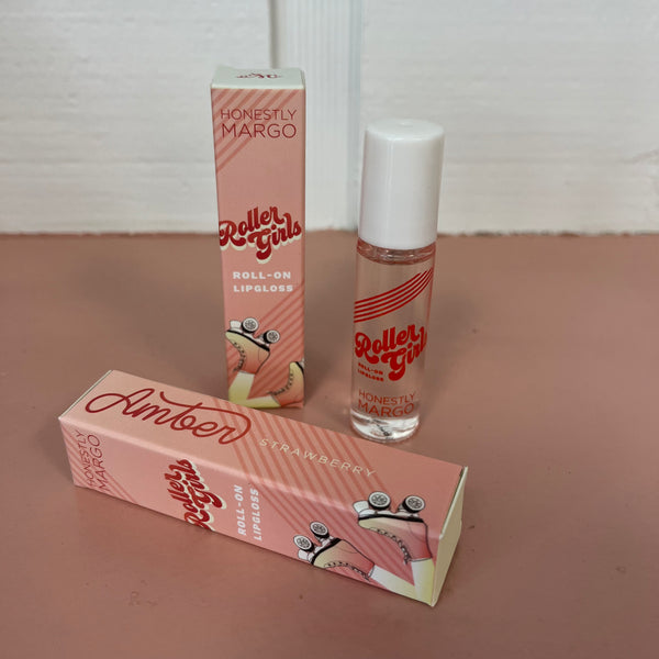 In the back there is the peach Honestly Margo Roller Girls Lip Gloss box next to the lip gloss. The box is peach and says, "Roller Girls, Roll-on Lipgloss", and the lip gloss is in a clear tube with a white cap and says, "Roller Girls" in red. In the front the box is laying on its side. On the top it says, "Amber, Strawberry", and on the side it says, "Roller Girls, Roll-on Lipgloss".