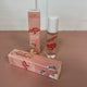 In the back there is the peach Honestly Margo Roller Girls Lip Gloss box next to the lip gloss. The box is peach and says, "Roller Girls, Roll-on Lipgloss", and the lip gloss is in a clear tube with a white cap and says, "Roller Girls" in red. In the front the box is laying on its side. On the top it says, "Amber, Strawberry", and on the side it says, "Roller Girls, Roll-on Lipgloss".