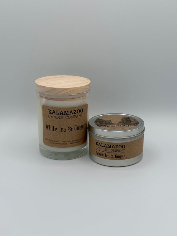 The White Tea and Ginger Kalamazoo Candle jar next to the White Tea and Ginger Kalamazoo Candle tin. The jar is clear through which you can see the white candle with a light wood lid and a brown paper label that reads, "Kalamazoo Candle Company, White Tea and Ginger". The tin is a small cylindrical metal tin with a brown paper label that says, "Kalamazoo Candle Company, White Tea and Ginger".