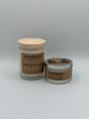 The White Tea and Ginger Kalamazoo Candle jar next to the White Tea and Ginger Kalamazoo Candle tin. The jar is clear through which you can see the white candle with a light wood lid and a brown paper label that reads, "Kalamazoo Candle Company, White Tea and Ginger". The tin is a small cylindrical metal tin with a brown paper label that says, "Kalamazoo Candle Company, White Tea and Ginger".